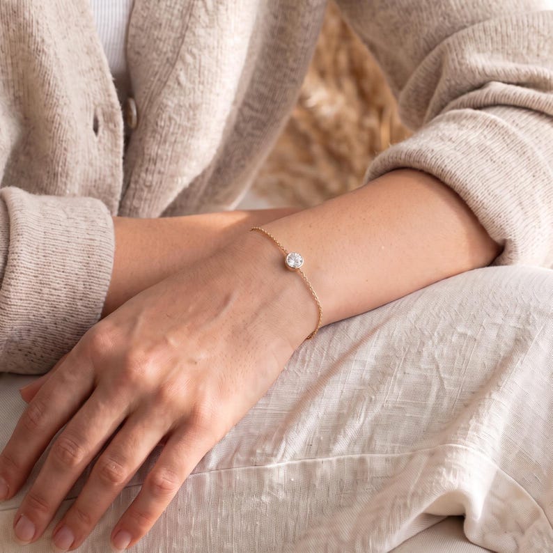May include: A delicate gold chain bracelet featuring a single, round, clear faceted stone set in a simple gold bezel. The bracelet rests on a wrist, showcasing a minimalist design with a thin, polished metal chain and a secure, centered gemstone.