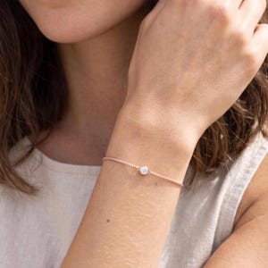May include: A delicate rose gold chain bracelet featuring a single, round, bezel-set clear gemstone at the center. The thin, twisted metal chain secures around the wrist with a minimalist design, highlighting the sparkling stone as the focal point.
