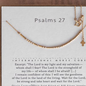 May include: A gold-toned chain necklace featuring a Morse code pattern of small gold and silver beads. The necklace includes a small cross charm and is displayed on a white card printed with the text Psalms 27 and the corresponding Morse code key.