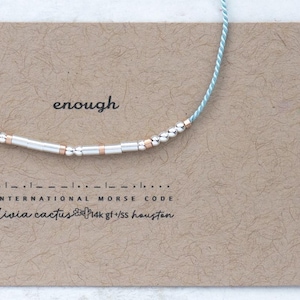 May include: A silver and rose gold beaded necklace with the word "enough" printed on a brown card. The card also includes International Morse Code and the text "olivia cactus". The necklace is strung on a light blue cord.