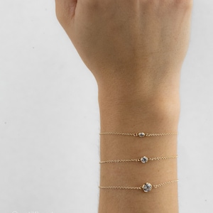 May include: Three delicate gold chain bracelets, each featuring a single round bezel-set clear stone. The bracelets are arranged vertically on a wrist, with the stone size increasing from the top bracelet to the bottom bracelet.