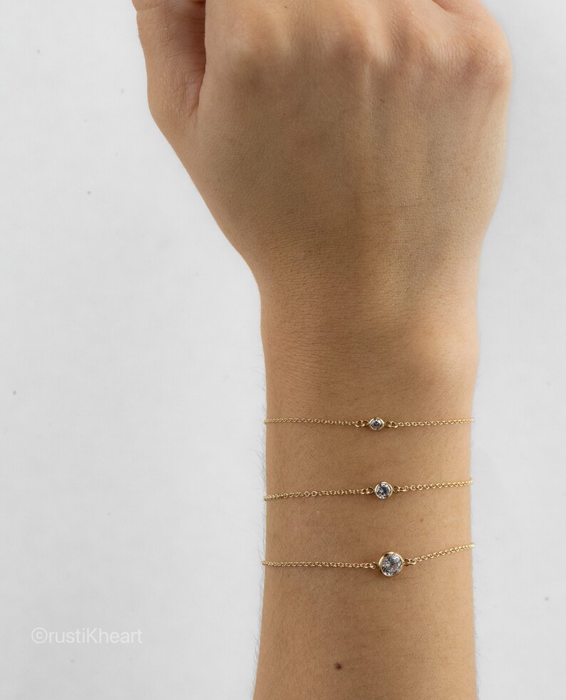 May include: Three delicate gold chain bracelets, each featuring a single round bezel-set clear stone. The bracelets are arranged vertically on a wrist, with the stone size increasing from the top bracelet to the bottom bracelet.