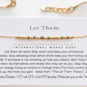 May include: Gold chain bracelet with small green, pink, and gold beads. The bracelet is on a white card with the text "Let Them." and "International Morse Code" in black. The text continues with a message about letting go of control and finding peace. The card also includes the brand name "Olivia Cactus" and the text "14k GF+/.925 88 Jewelry Made for you in the USA."