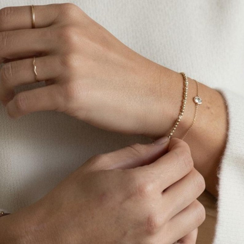 May include: Two gold rings with thin bands worn on the fingers and two gold bracelets on the wrist. One bracelet features a delicate chain with a single small round clear stone, while the other is a gold beaded chain bracelet.