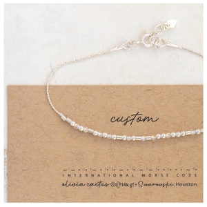 May include: A delicate silver bracelet with a beaded design, presented on a brown card. The card features the word "custom" in cursive, along with text in Morse code and the brand name "olivia cactus".