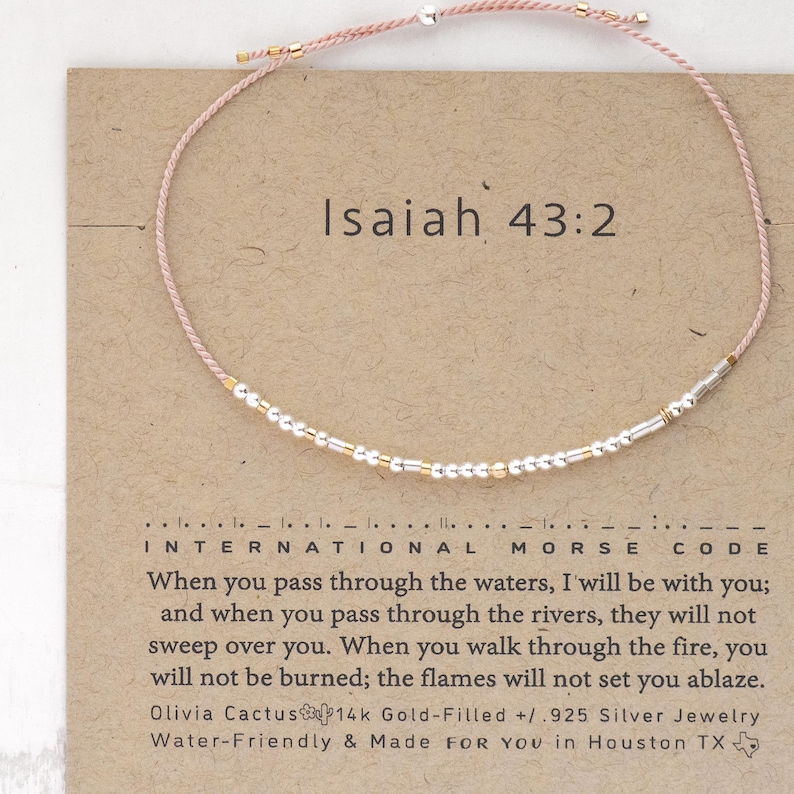 May include: A pink string bracelet with silver and gold beads. The bracelet has the text "Isaiah 43:2" on a card and the words "International Morse Code" below it. The card also includes a quote from Isaiah 43:2.