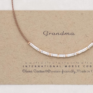 May include: A necklace with silver and gold beads spelling "Grandma" in Morse code. The necklace is on a brown card with the words "Grandma" and "International Morse Code" printed on it. The card also has the text "Olivia Cactus" and "water-friendly, Made in TX".