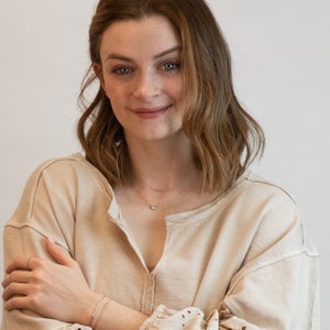 May include: A woman wearing a light beige sweater with a delicate gold necklace and bracelet. The necklace features a small pendant.