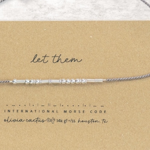 May include: A silver Morse code bracelet with the phrase "let them" spelled out in beads. The bracelet is on a tan card with the text "INTERNATIONAL MORSE CODE" and "olivia cactus 14k gf+/ss houston. Tx".