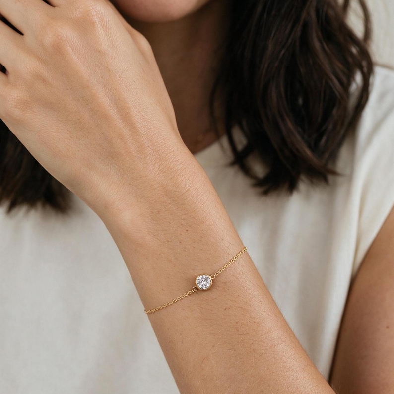 May include: A delicate gold chain bracelet featuring a single round, clear, faceted stone set in a circular gold bezel. The bracelet is secured with a thin, polished gold link chain that wraps around the wrist, showcasing a minimalist design.