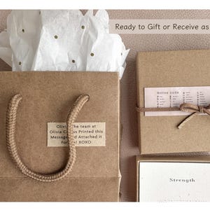 May include: A brown paper gift bag with a rope handle. The bag has a small white label with the text "Olivi the team at Olivia Cus Printed this Messaged Attached it For! XOXO". The bag is sitting on a white tissue paper with gold glitter. The bag is next to a brown box with a white label that says "morse code" and a brown box with a white label that says "Strength".