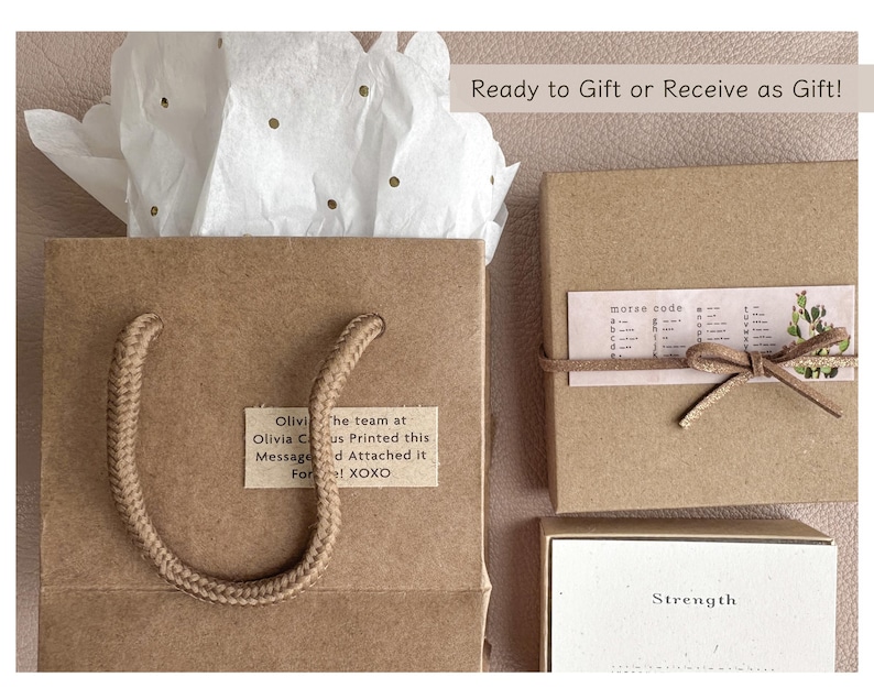 May include: A brown paper gift bag with a rope handle. The bag has a small white label with the text "Olivi the team at Olivia Cus Printed this Messaged Attached it For! XOXO". The bag is sitting on a white tissue paper with gold glitter. The bag is next to a brown box with a white label that says "morse code" and a brown box with a white label that says "Strength".