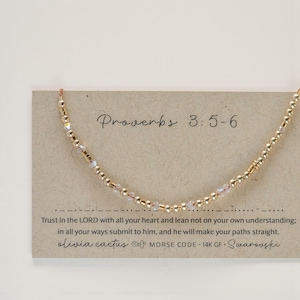May include: A gold-colored beaded necklace with a delicate design. The necklace is displayed on a card with the text "Proverbs 3:5-6" and a quote. The card also includes the text "olivia cactus MORSE CODE - 14K GF + Swarovski."