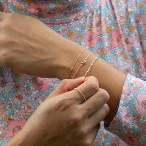 May include: Three delicate gold-toned bracelets worn on the wrist, including one with a small clear stone, and a matching gold-toned beaded ring on the finger. The jewelry features fine chains and small metallic spheres, creating a layered look.