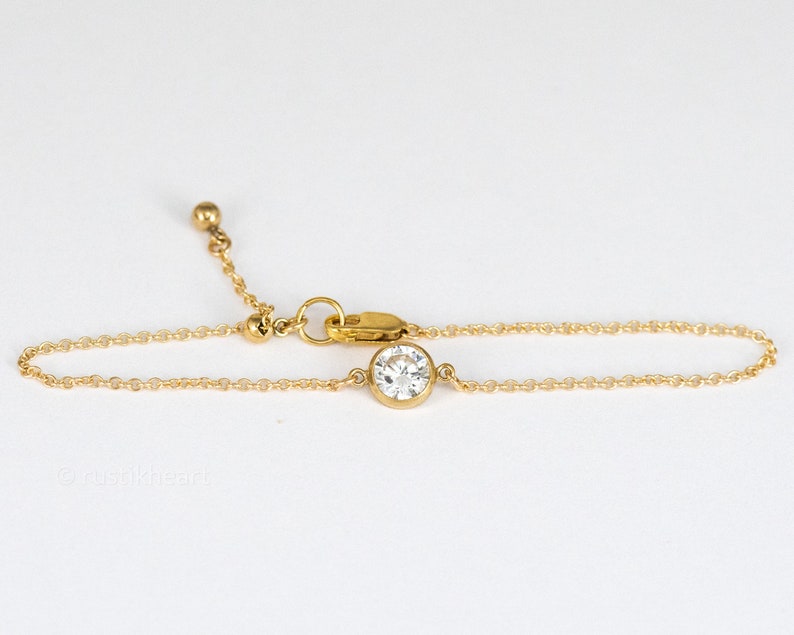 May include: A gold-toned chain bracelet featuring a single round, clear, faceted stone set in a bezel setting. The bracelet includes a delicate link chain, a lobster claw clasp, and an adjustable extension chain finished with a small gold-toned bead.