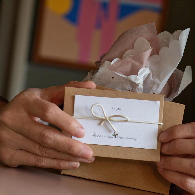 May include: A brown cardboard gift box topped with white and dusty pink scalloped tissue paper. A white rectangular card is tied to the front with thin white string in a bow, featuring the text for you and olivia cactus Morse-dainty.
