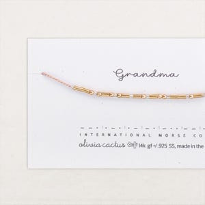 May include: A gold beaded necklace spelling "Grandma" in Morse code, presented on a white card. The card also displays the brand name "olivia cactus" and product details.