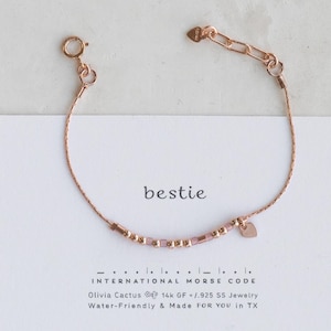 May include: A rose gold bracelet with a heart charm and a beaded section that spells out "bestie" in international morse code. The bracelet is on a white card with the text "bestie", "INTERNATIONAL MORSE CODE", "Olivia Cactus 14k GF/.925 SS Jewelry", and "Water-Friendly & Made FOR YOU in TX".
