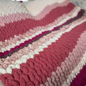 May include: A handmade, chunky knit blanket in shades of pink, white, and burgundy. The blanket features a striped pattern with varying widths of color blocks. The soft, textured yarn creates a cozy and inviting aesthetic, perfect for home decor.