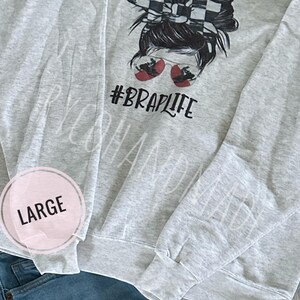 May include: A light gray sweatshirt with a graphic of a woman with a checkered visor and sunglasses. The text "#BRAPLIFE" is below the graphic. The word "LARGE" is in a pink circle. The sweatshirt is paired with blue jeans.