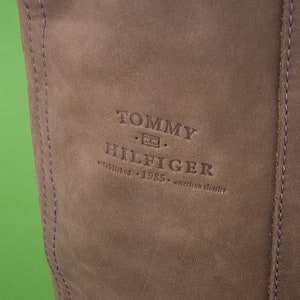 May include: Close-up of a brown leather bag with the Tommy Hilfiger logo embossed in the center. The logo reads "Tommy Hilfiger established 1985 american classics."