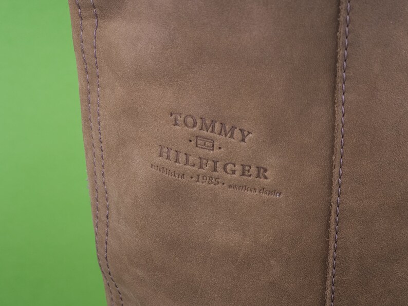 May include: Close-up of a brown leather bag with the Tommy Hilfiger logo embossed in the center. The logo reads "Tommy Hilfiger established 1985 american classics."