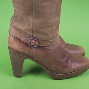 May include: Brown leather heeled boots with a buckle strap. The boots have a chunky heel and a rounded toe.