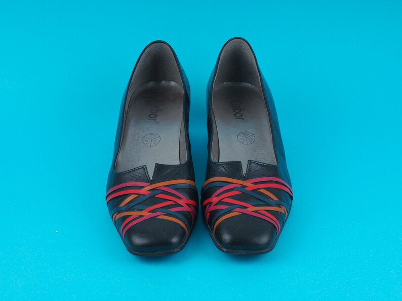 May include: A pair of black leather shoes with colorful woven straps. The shoes have a pointed toe and a low heel.