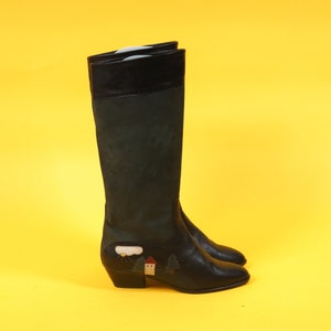 May include: Black suede knee-high boots with a small embroidered scene of a house and trees. The boots have a block heel.