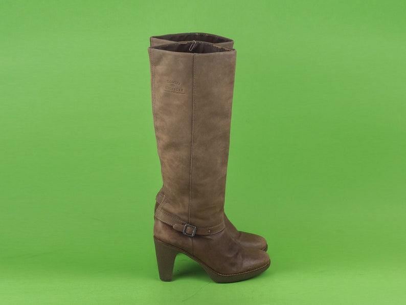 May include: Brown leather knee-high boots with a buckle detail and a block heel. The boots have the Tommy Hilfiger logo on the side.