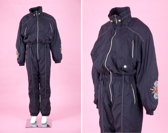 May include: A black ski suit with gold accents and embroidered designs. The suit has a zip closure and multiple pockets.
