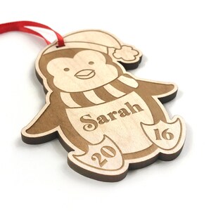 May include: A wooden penguin Christmas ornament with a red ribbon, a white hat, and a striped scarf. The ornament is personalized with the name "Sarah" and the year "2016".