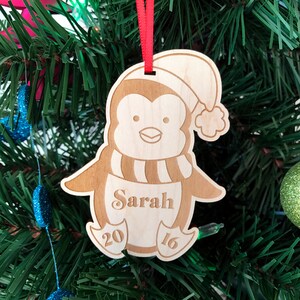 May include: A wooden penguin Christmas ornament with a red ribbon, a Santa hat, and the name "Sarah" engraved on the front. The ornament also has the year "2016" engraved on the bottom.