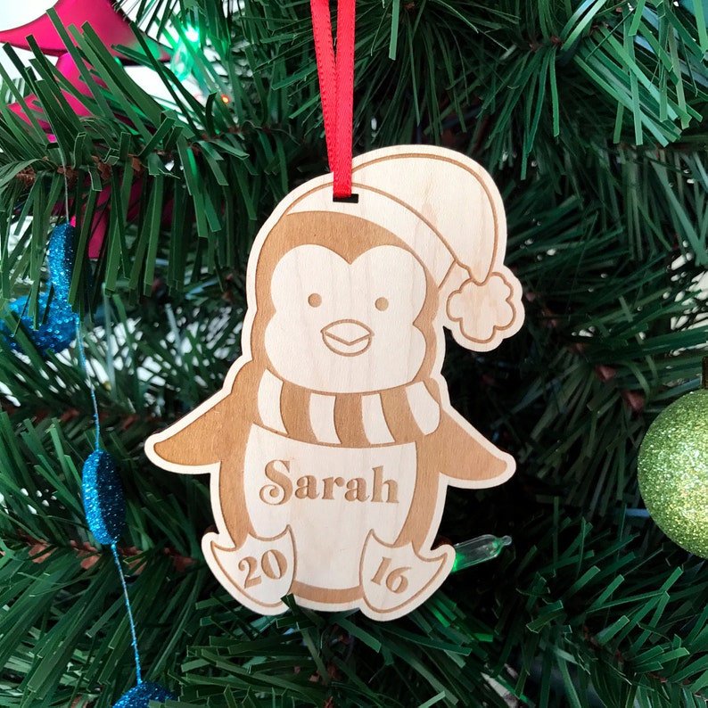 May include: A wooden penguin Christmas ornament with a red ribbon, a Santa hat, and the name "Sarah" engraved on the front. The ornament also has the year "2016" engraved on the bottom.