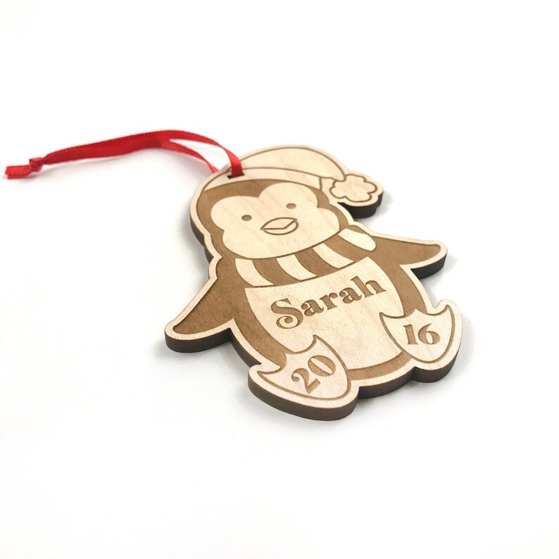 May include: A wooden penguin Christmas ornament with a red ribbon, a white hat, and a striped scarf. The ornament is personalized with the name "Sarah" and the year "2016".