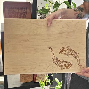 Op de afbeelding: Een licht houten snijplank met twee koi-karpers gegraveerd in het hout. De vissen kijken elkaar aan en bevinden zich in een yin-yang-positie.