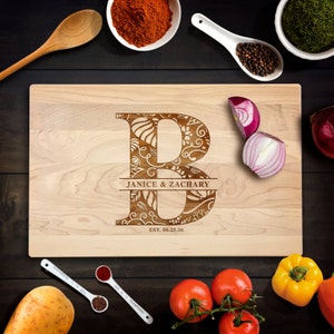Peut inclure: Une planche à découper en bois d'érable personnalisée avec une lettre B gravée décorative, les noms Janice & Zachary et la date 06.25.16. La planche est entourée de divers ingrédients de cuisine, notamment des tomates, des oignons et des épices.