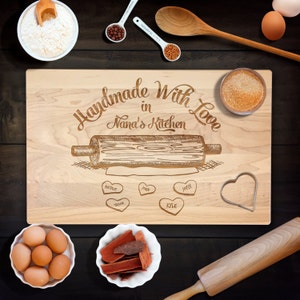 May include: A wooden chopping board with the text "Handmade With Love in Nana's Kitchen" engraved on it. The board also features a drawing of a rolling pin and a heart-shaped cookie cutter. There are five heart-shaped spaces for names, with the names "Natalie", "John", "Joyce", "Trevor", and "Kyle" engraved in them.