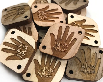 May include: A pile of wooden tags with a handprint and the words "HAND MADE" engraved on them. The tags are a light brown colour with a darker brown handprint.