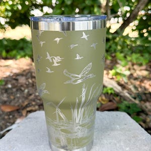 May include: An olive green stainless steel tumbler with a silver lid. The tumbler features a detailed etched design of flying ducks, reeds, and water. The design wraps around the entire tumbler, creating a nature-themed aesthetic.