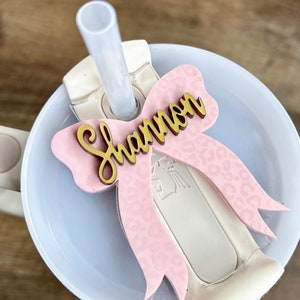 May include: A pink and gold bow with a leopard print design and the name "Shannon" written in gold letters. The bow is attached to a white tumbler with a straw.