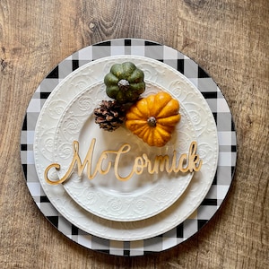 May include: A white plate with a black and white buffalo plaid charger. The plate has a decorative embossed design. There are three small gourds and a pine cone on the plate. A wooden name card with the name "McCormick" is placed on the plate.