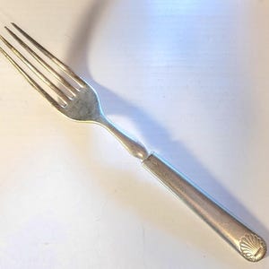 May include: A silver fork with four tines, resting on a white surface. The handle is rectangular with a seashell design at the end. The fork is made of metal and appears to be vintage.