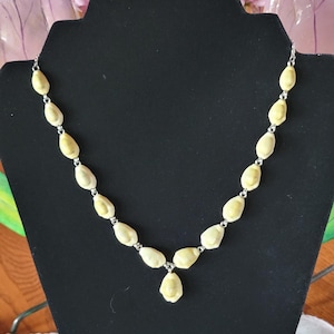 May include: A cream-colored necklace with a silver chain and pendant. The necklace features oval-shaped beads connected by small silver links. The pendant is a single, teardrop-shaped bead. The necklace is displayed on a black jewelry stand.