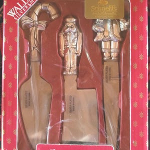 May include: A 3-piece cheese set in a red box. The set includes three silver-plated cheese knives with decorative handles: a candy cane, a nutcracker, and Santa Claus. The box has the text "3-Piece Cheese Set".