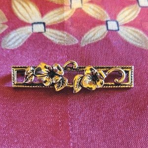 May include: A gold-toned metal bar pin featuring two detailed flower blossoms with leaves. The flowers are centered within a rectangular frame with decorative flourishes. The pin is likely a vintage or antique jewelry piece.