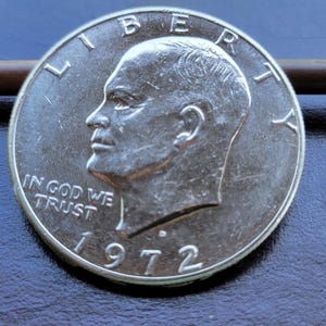 Op de afbeelding: Een zilveren 1972 Eisenhower dollar munt. De munt toont een profiel van president Dwight D. Eisenhower en de inscriptie "IN GOD WE TRUST". Het jaartal "1972" is prominent aanwezig. De munt ligt op een donkerblauwe ondergrond.