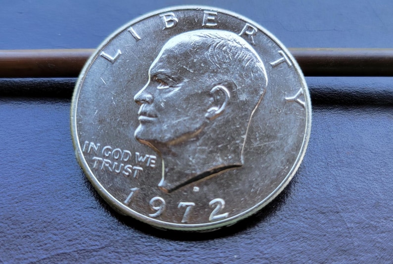 Op de afbeelding: Een zilveren 1972 Eisenhower dollar munt. De munt toont een profiel van president Dwight D. Eisenhower en de inscriptie "IN GOD WE TRUST". Het jaartal "1972" is prominent aanwezig. De munt ligt op een donkerblauwe ondergrond.