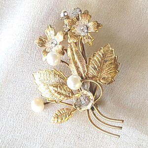 May include: A gold-toned floral brooch featuring detailed leaves, flowers with clear crystal accents, and faux pearls. The brooch has a classic design, perfect for adding a touch of elegance to any outfit.
