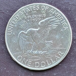 Op de afbeelding: Een zilveren dollar munt met een adelaar met uitgespreide vleugels, omringd door de woorden "UNITED STATES OF AMERICA" en "ONE DOLLAR". De munt heeft ook de inscriptie "E PLURIBUS UNUM".